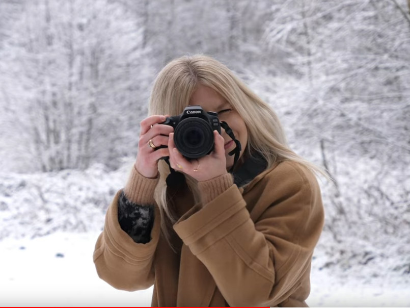  Blondynka trzyma aparat fotograficzny przy twarzy, wykonuje zdjęcie. W tle zimowa sceneria, zaśnieżone drzewa. 